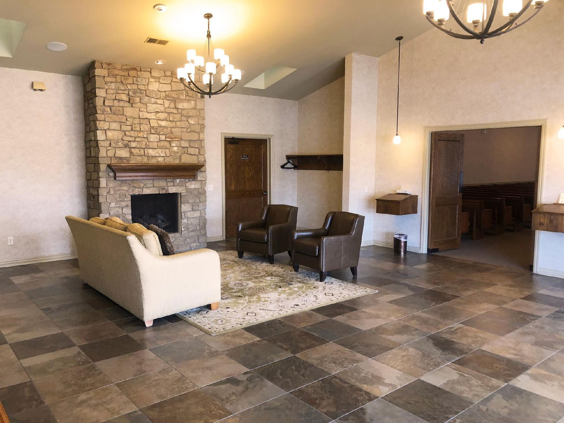 Spacious living room with stone fireplace, beige chairs, and tiled floor under chandeliers