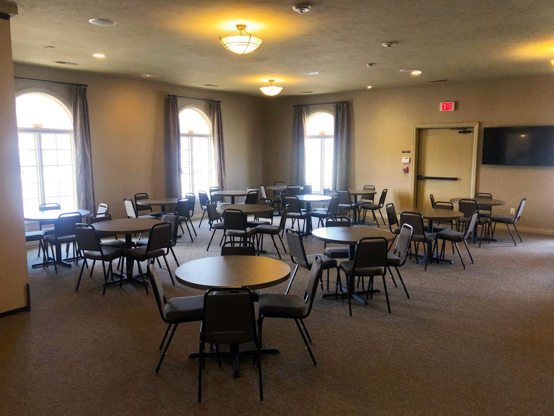 Empty banquet hall with round tables, black chairs, arched windows, and a lit exit sign by the door