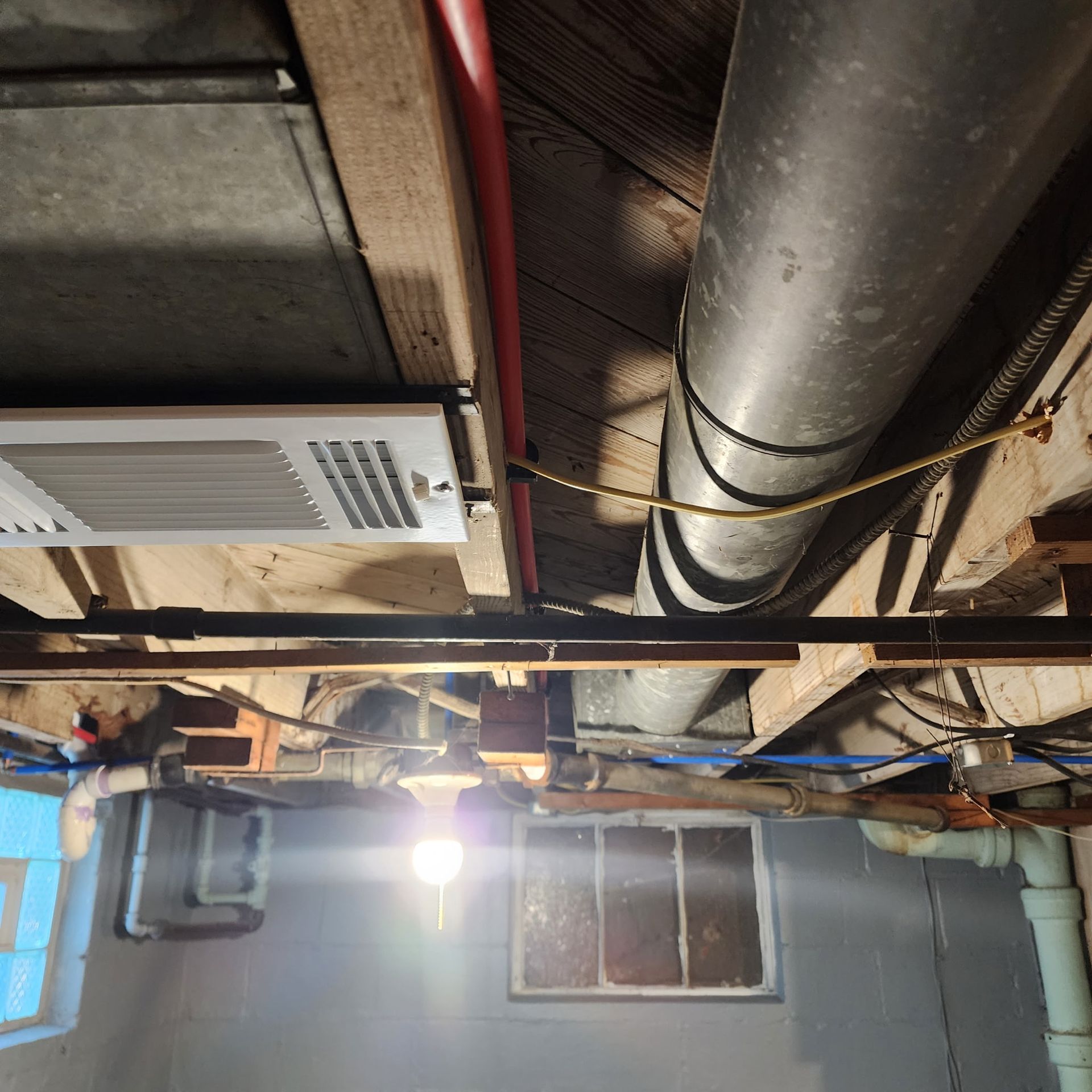 A basement ceiling with a white vent, a large metal duct, exposed wooden beams, and a hanging lightbulb.
