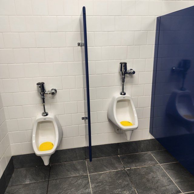 Two white porcelain urinals with yellow drain covers mounted on a white tiled wall, separated by a dark blue partition.