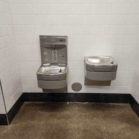 Wall-mounted metal water bottle filling station and a lower drinking fountain on a white tiled wall.