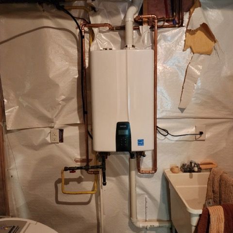 A wall-mounted white tankless water heater with copper piping, installed in a basement near a utility sink.