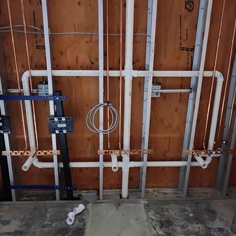 Plumbing and electrical rough-in installation featuring PVC pipes, copper lines, and metal studs on a plywood wall.