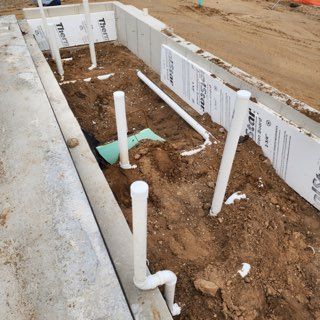 Construction site view of vertical white PVC plumbing pipes installed in a trench within a concrete foundation.