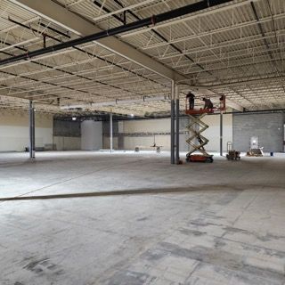 A large, empty warehouse interior with exposed ceilings, concrete floors, and workers on a scissor lift.