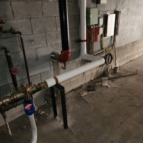A basement wall with exposed black, red, and white piping and electrical components mounted on gray concrete blocks.