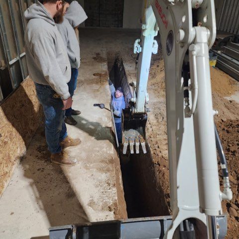 Two people stand on a concrete floor watching a third person standing inside a narrow trench next to an excavator.