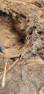 A metal pipe protrudes from the dirt wall of an excavated trench, with a shovel handle visible in the foreground.
