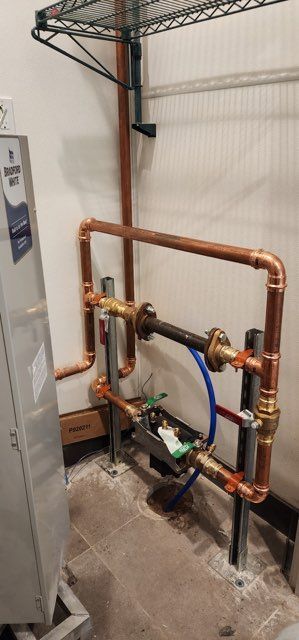 A water meter assembly with copper pipes and a flanged iron pipe section mounted against a wall in a utility area.