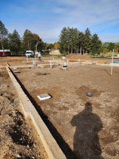 A residential construction site showing a concrete foundation perimeter and various pipe stubs emerging from dirt.