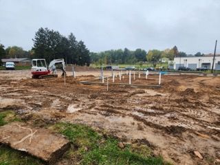 A construction site with a white mini-excavator and vertical white pipes protruding from a dirt foundation area.