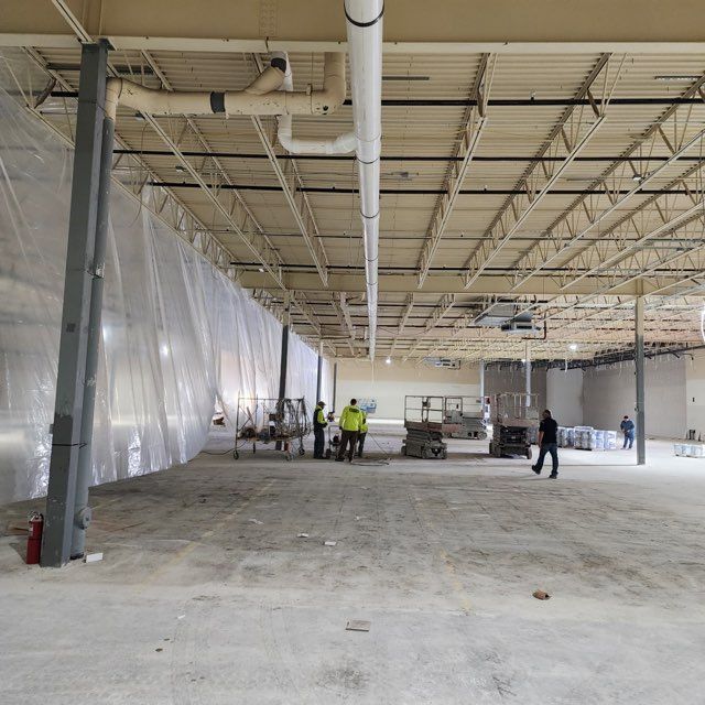 Construction workers stand inside a large, open-plan commercial space undergoing renovation with plastic sheeting dividers.