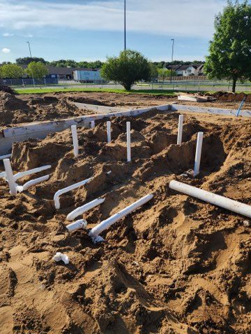 A construction site foundation with white PVC plumbing pipes sticking out of brown dirt in a sunny outdoor setting.
