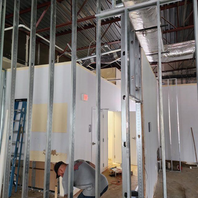 A worker assembles metal framing for a wall inside a building under construction with exposed ceilings and interior doors.