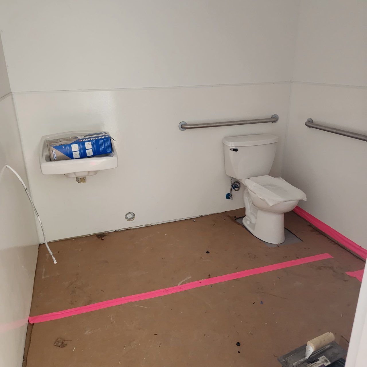 A public restroom under construction with a toilet, sink, and grab bars on the walls, featuring pink floor marking tape.