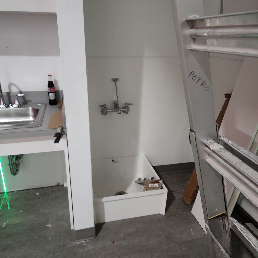 A utility sink with faucet, pipes, and a partial counter in an unfinished, white-walled room under construction.