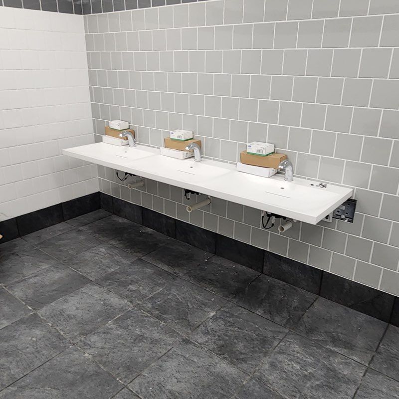 A wall-mounted white vanity with three sinks, chrome faucets, and boxes of soap, set against light grey tiled walls.