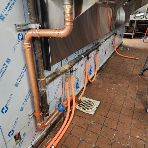 Commercial kitchen wall with copper pipes and orange gas hoses connected to a main black pipe over a tiled floor.