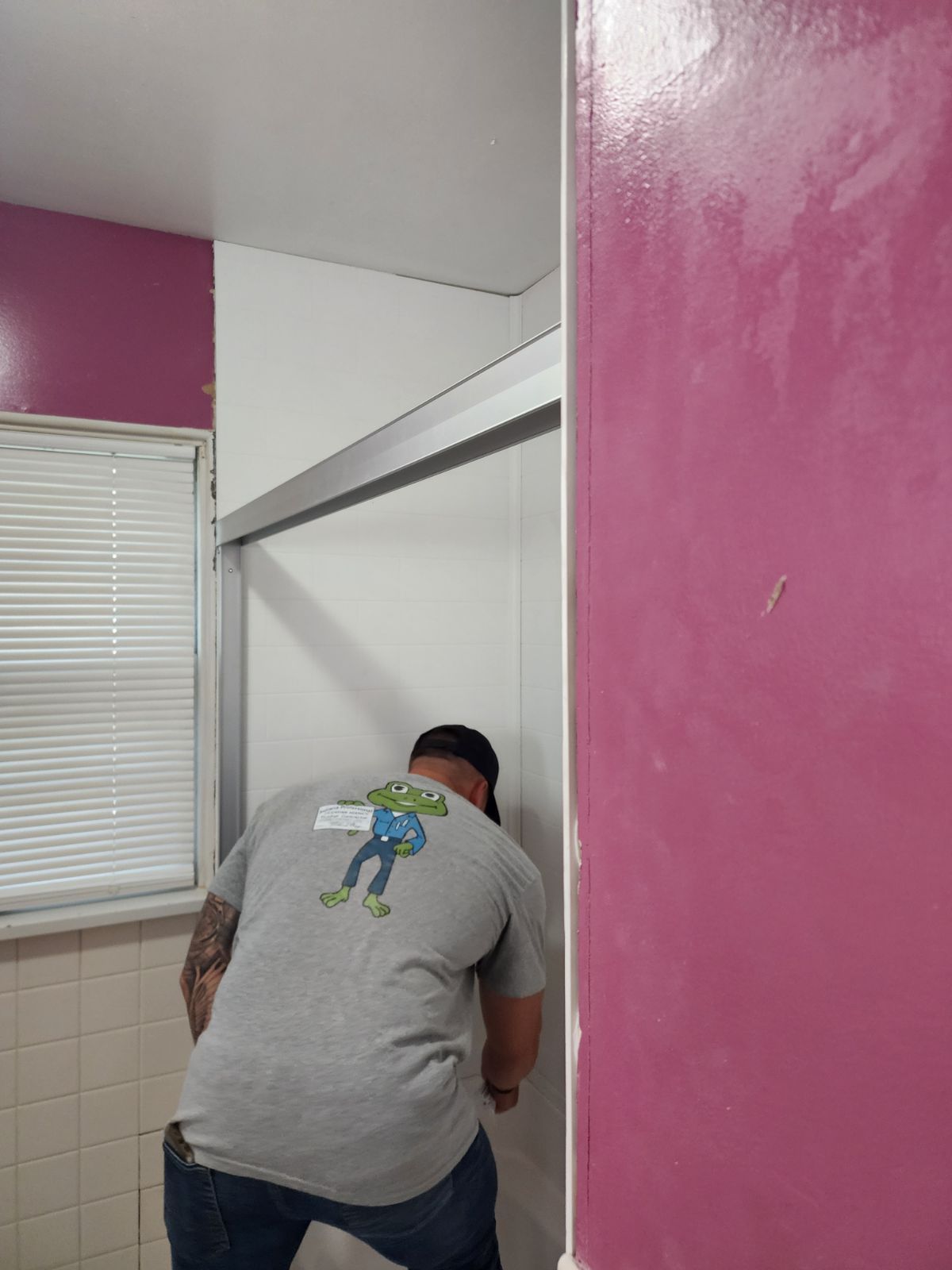A person in a gray graphic t-shirt works to install a silver metal frame inside a bathroom shower area.