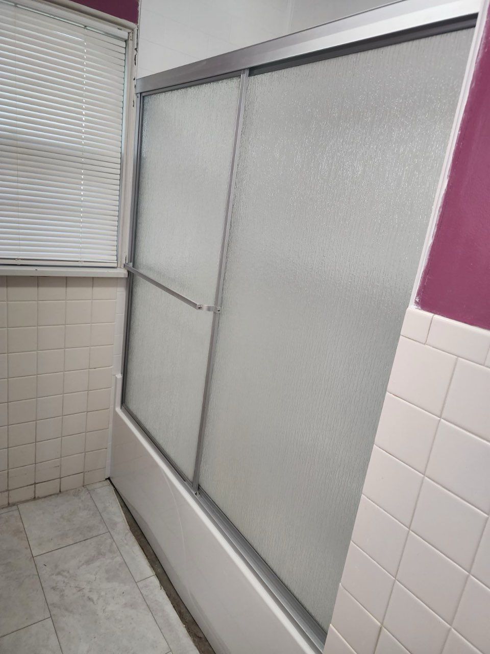 A bathroom featuring a shower with sliding, textured glass doors, tiled walls, and a window with blinds.