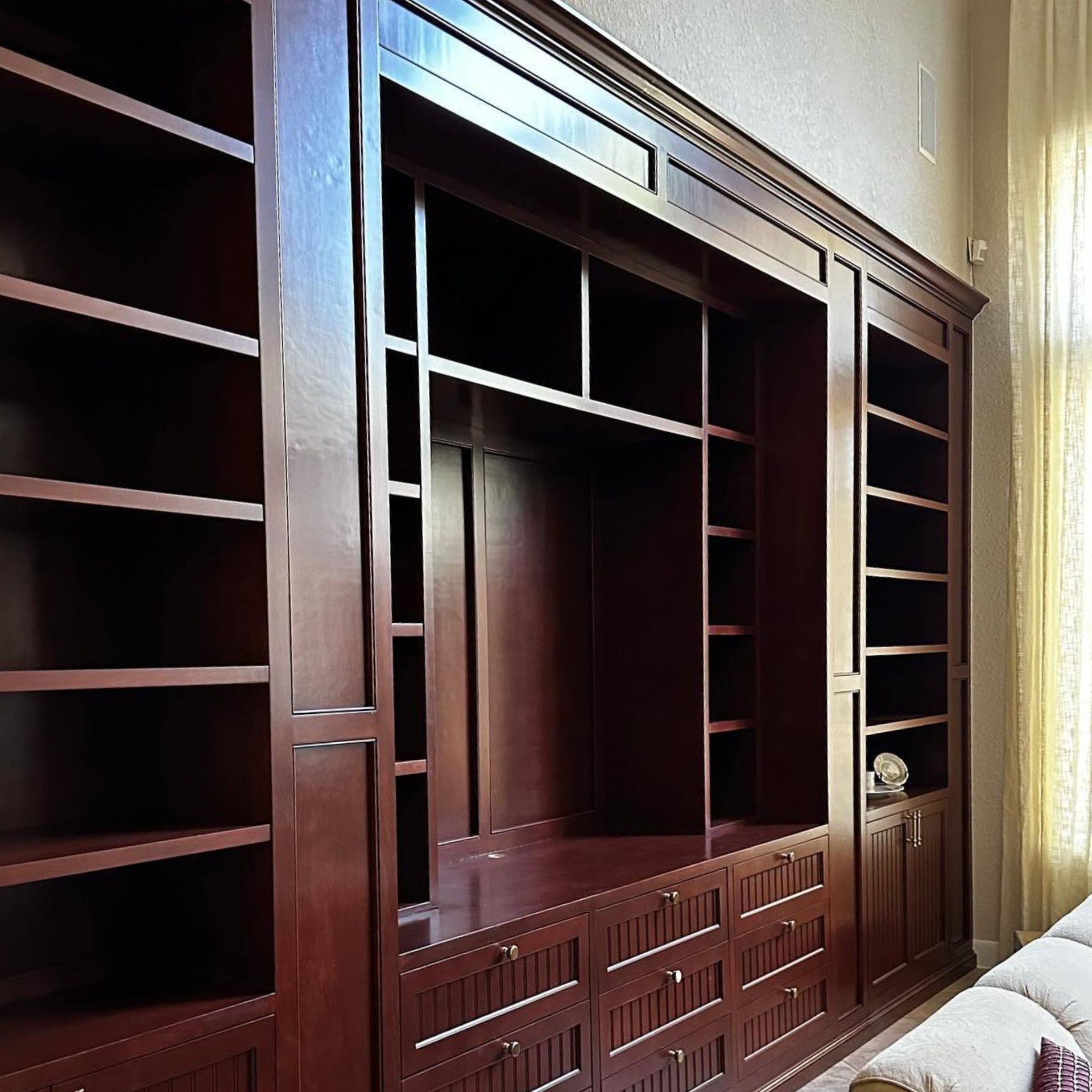 Dark wooden built-in shelving and cabinets in a bright room beside a curtained window.