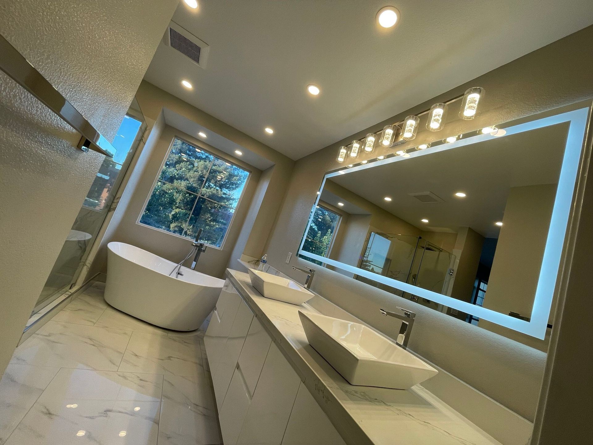 Luxury master bathroom remodel featuring a freestanding soaking tub by a large picture window, dual vessel sinks on a sleek white vanity, illuminated LED backlit mirror, recessed lighting, marble-look porcelain flooring, and frameless glass shower — custom design by Dream Build Services in the Bay Area.