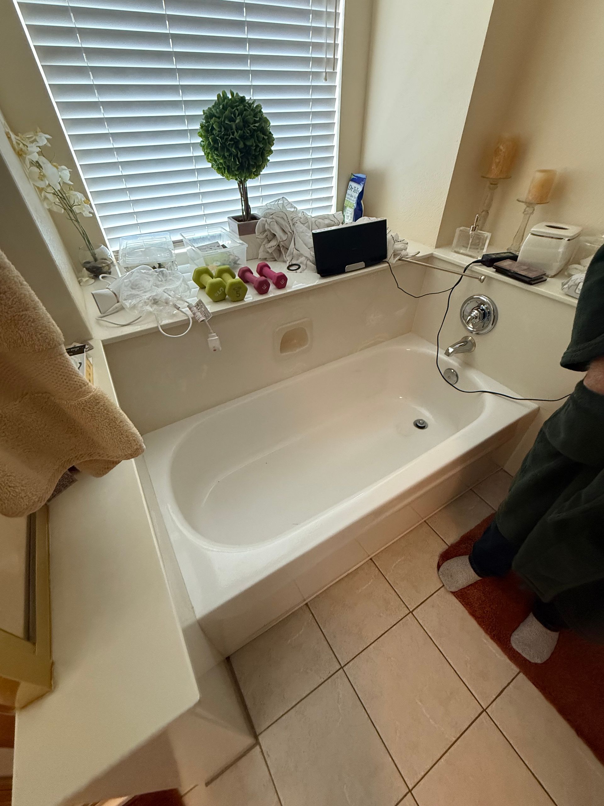 Bathtub beneath a window with blinds, decorated with plants, toiletries, and a shower hose.