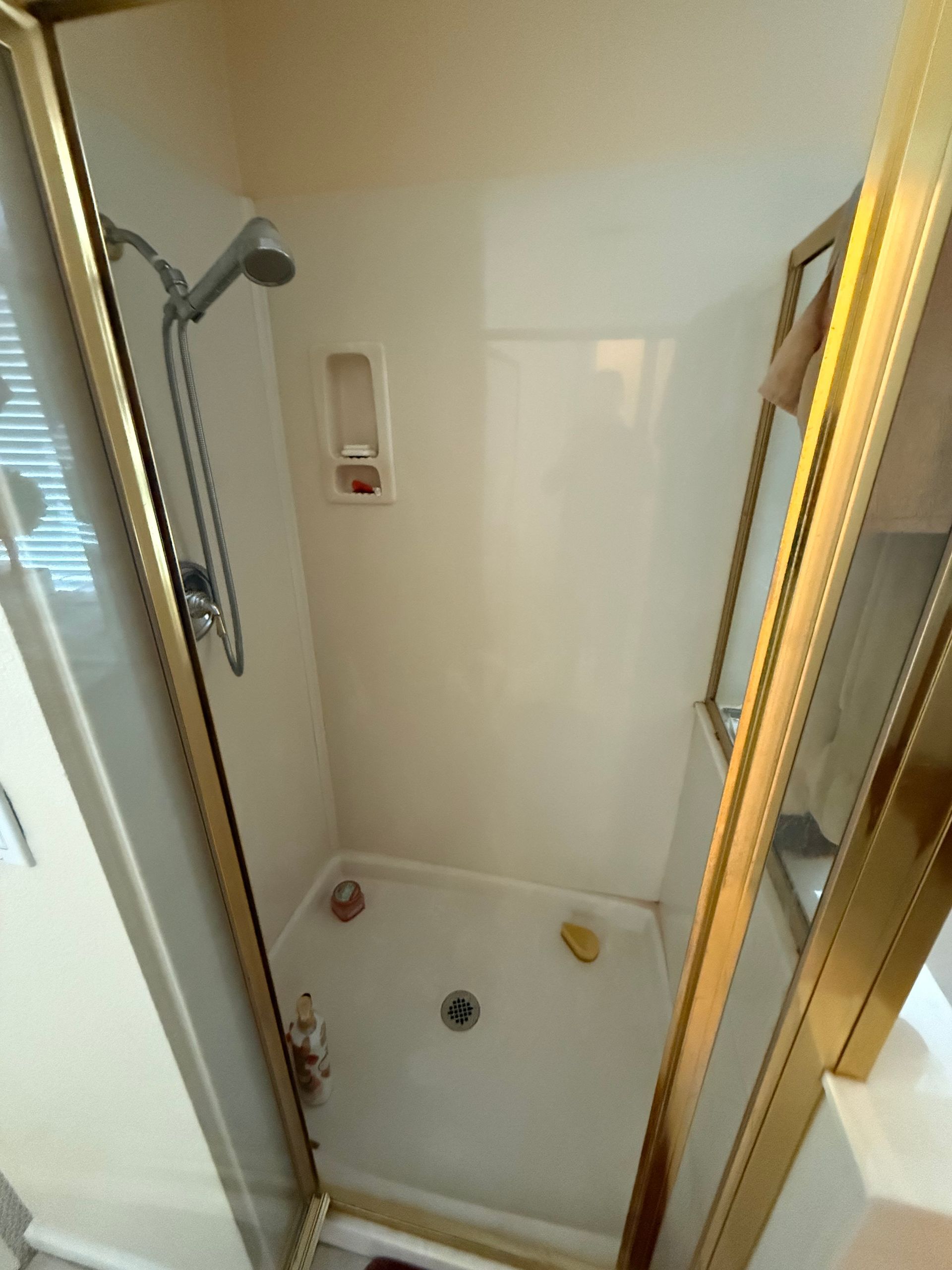 Empty shower stall with white walls, handheld showerhead, and gold-framed glass door.