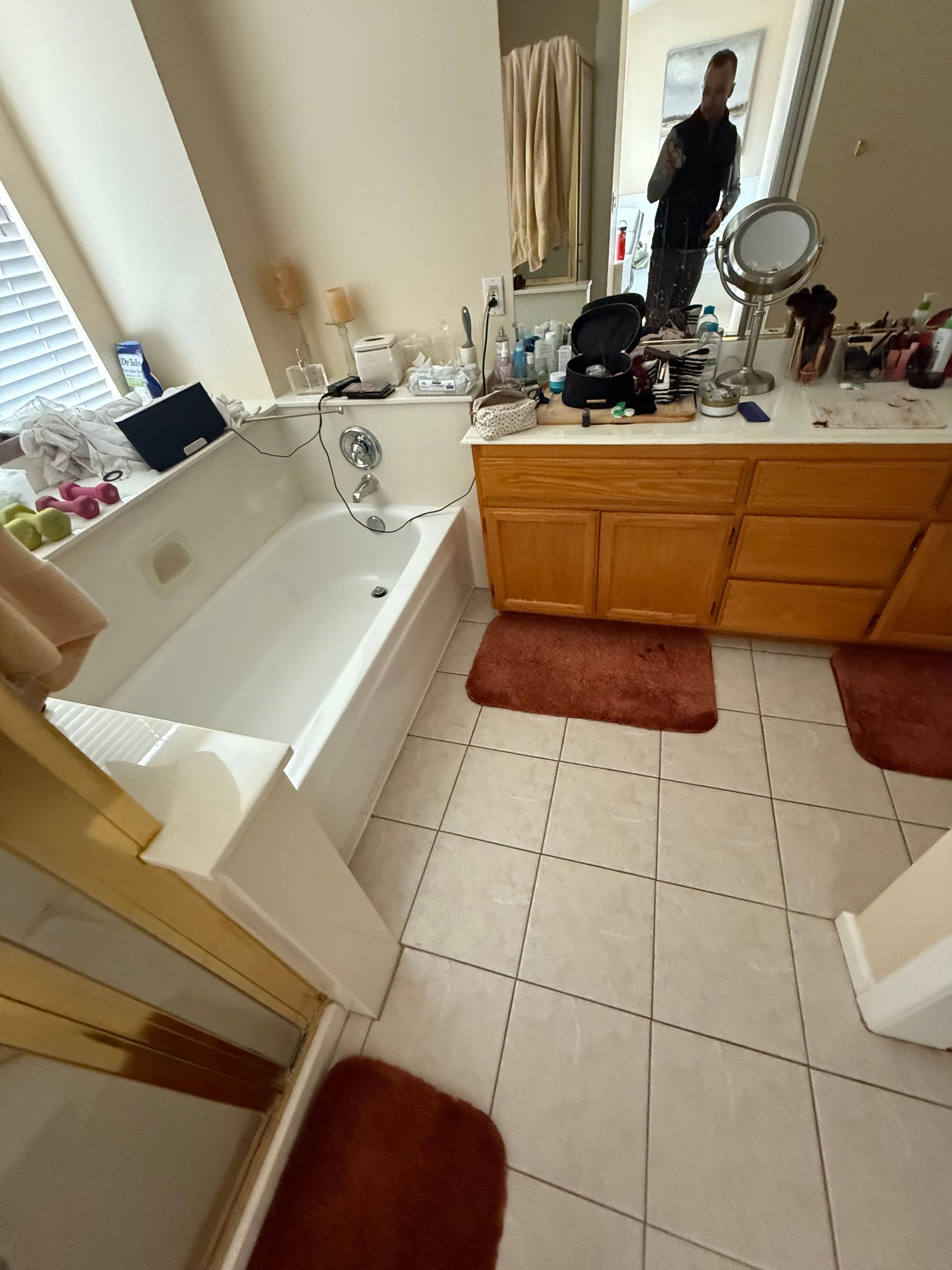Bathroom with white tub, wooden vanity, red rugs, and a person reflected in the mirror.
