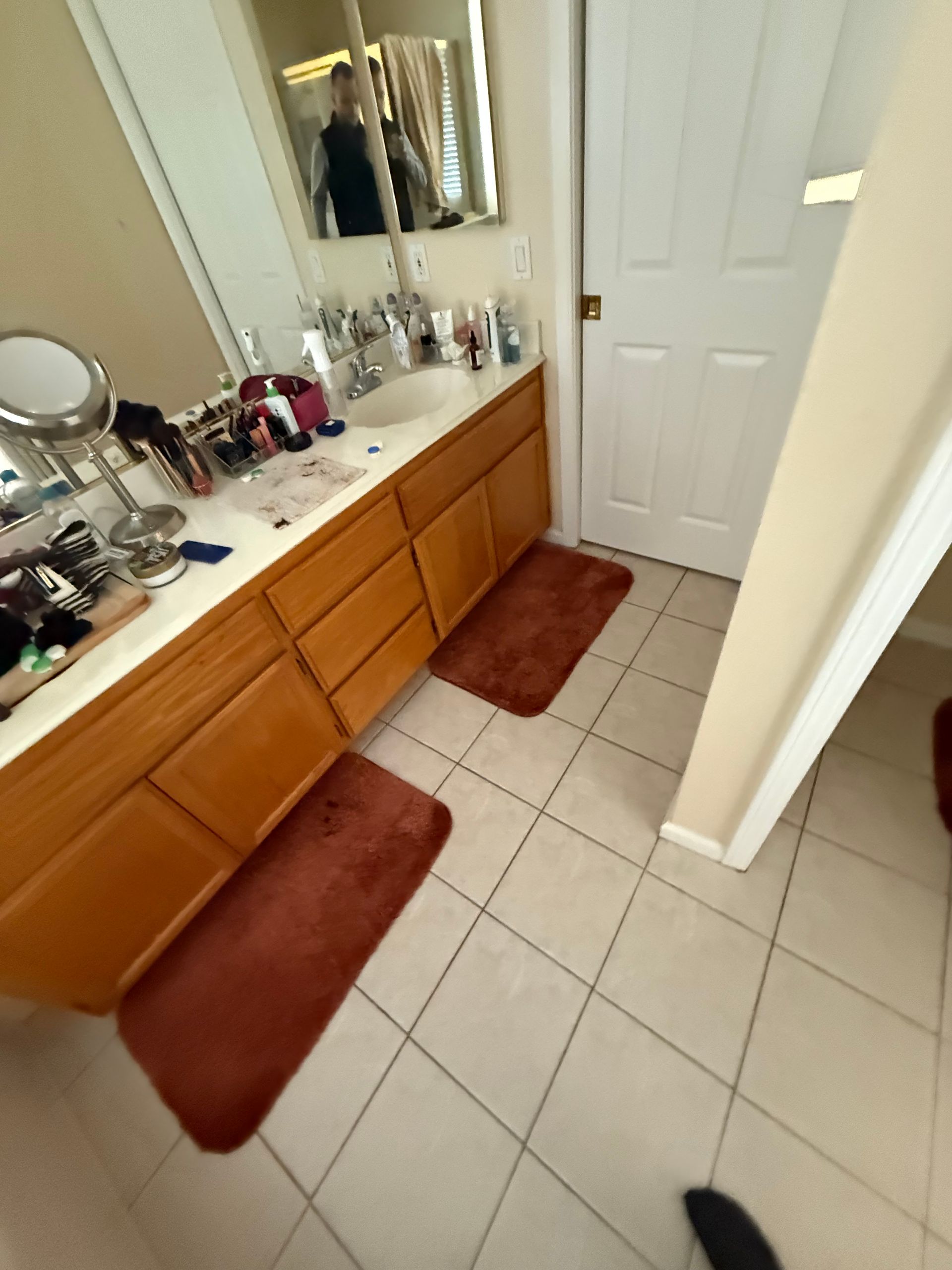 Bathroom vanity with mirror, sink, countertop items, and brown rugs on tiled floor.