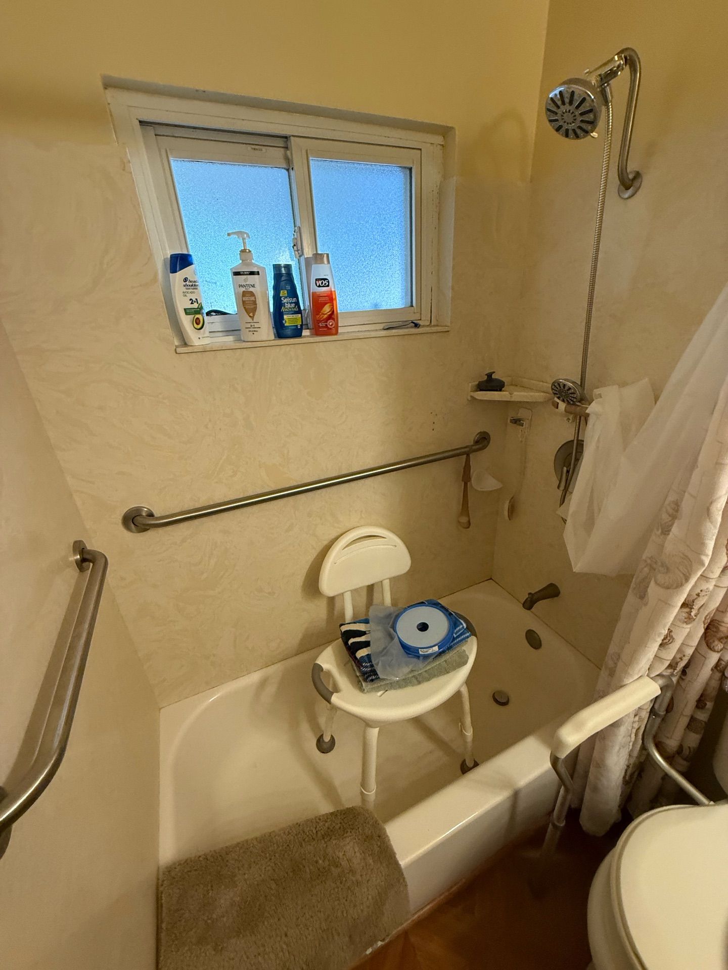 Bathroom with a tub, grab bars, shower chair, and toiletries on a window ledge.