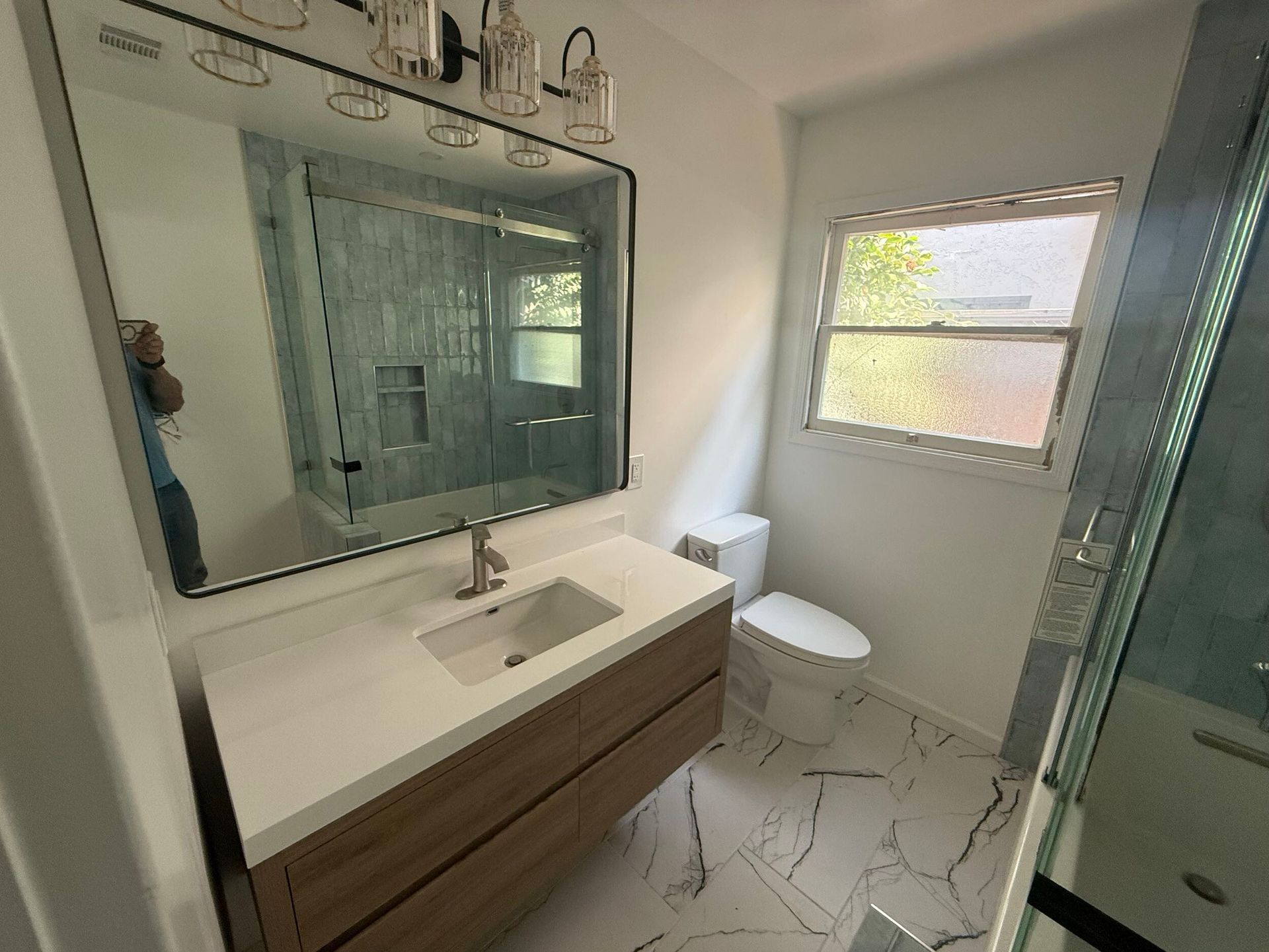Modern bathroom remodel featuring a sleek floating vanity with quartz countertop, frameless LED mirror, and upgraded lighting fixtures. The shower area is designed with glass doors, rainfall showerhead, and elegant blue ceramic wall tiles for a spa-like feel. White marble-look porcelain floor tiles add a touch of luxury and durability. Perfect blend of contemporary design and functionality, ideal for homeowners in the Bay Area looking for stylish bathroom renovation ideas.