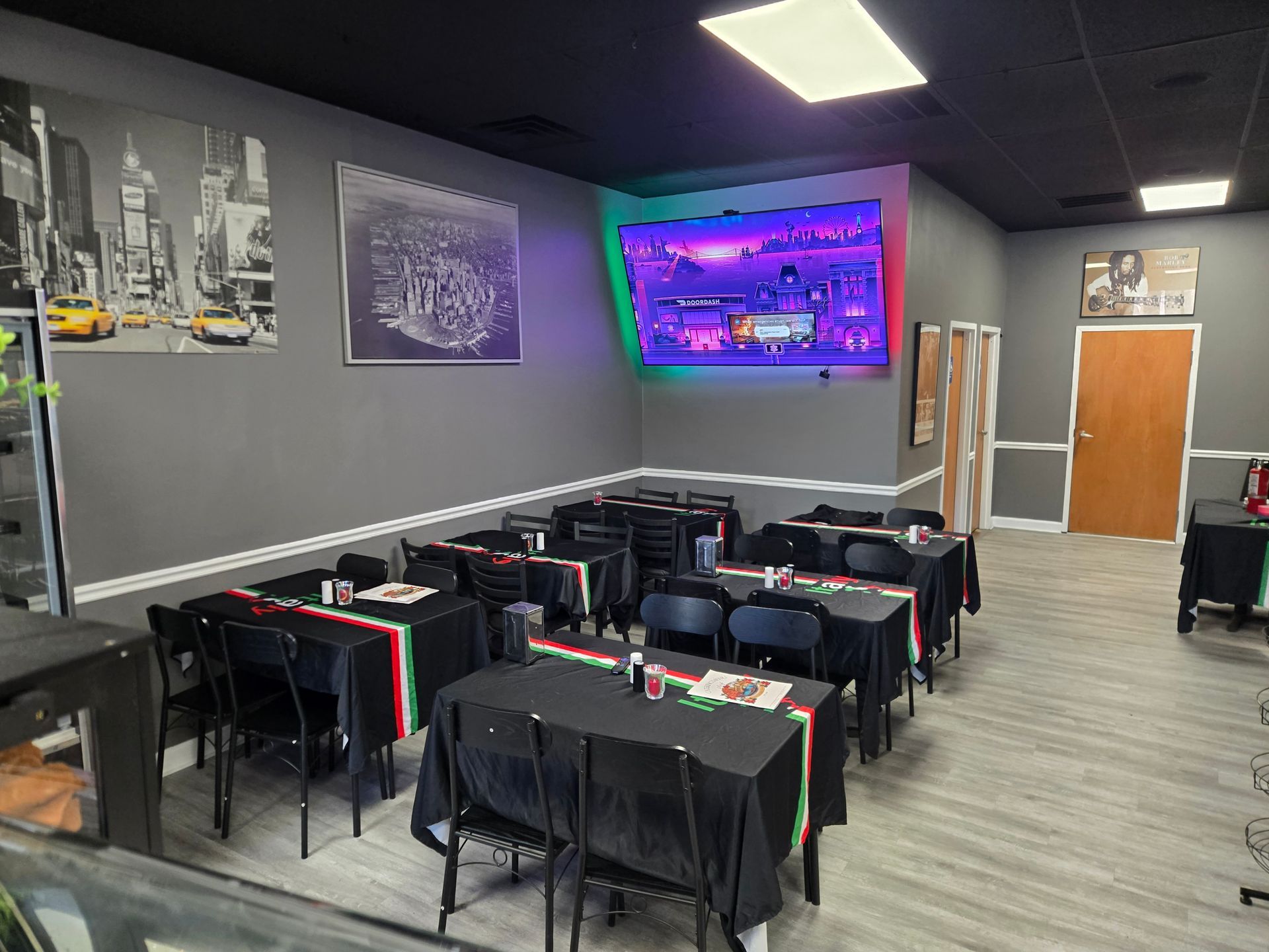 Empty restaurant dining room with black tables and red-trimmed chairs, wall art, and a TV on the back wall