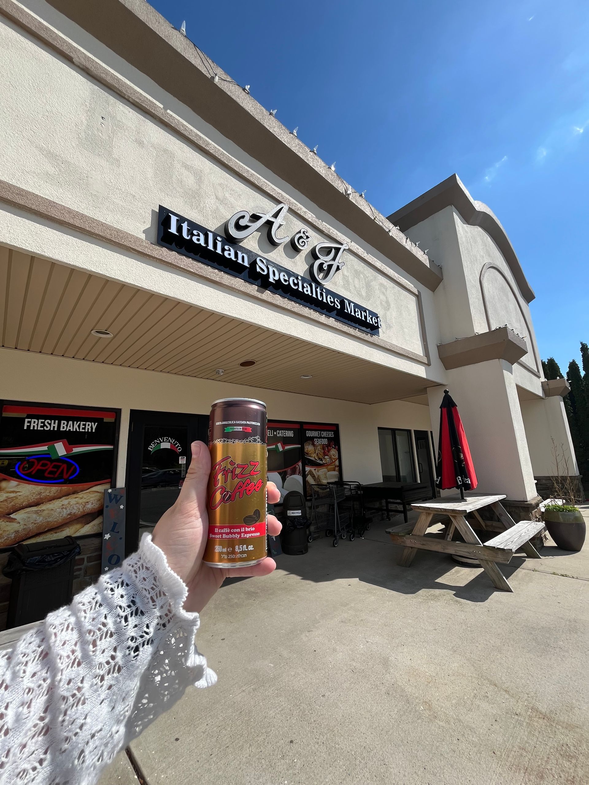 Hand holding a can outside a storefront with a blue sky and outdoor seating.