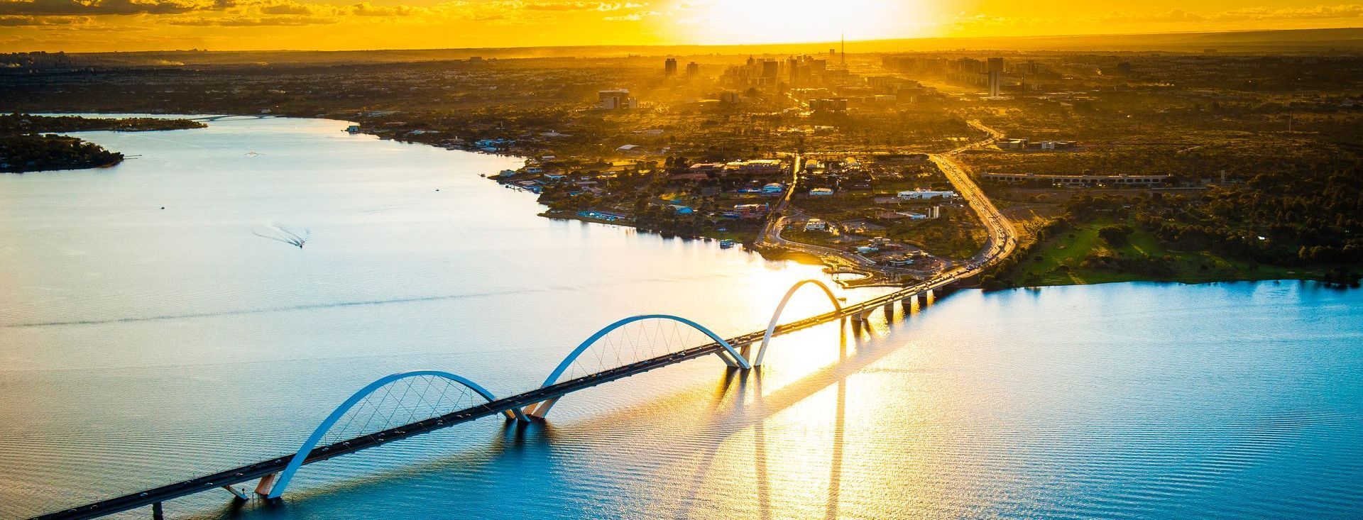 Uma ponte com arcos azuis sobre um lago, com a cidade ao fundo. Luz do sol dourada e reflexos na água.
