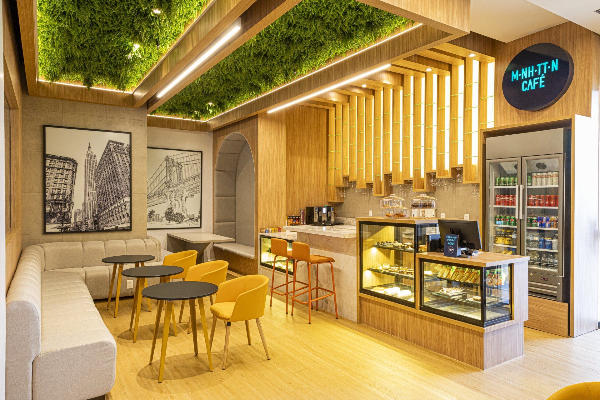 Interior do café com detalhes em madeira, mesas pequenas e uma vitrine. Também são exibidas plantas e uma arte urbana em preto e branco.