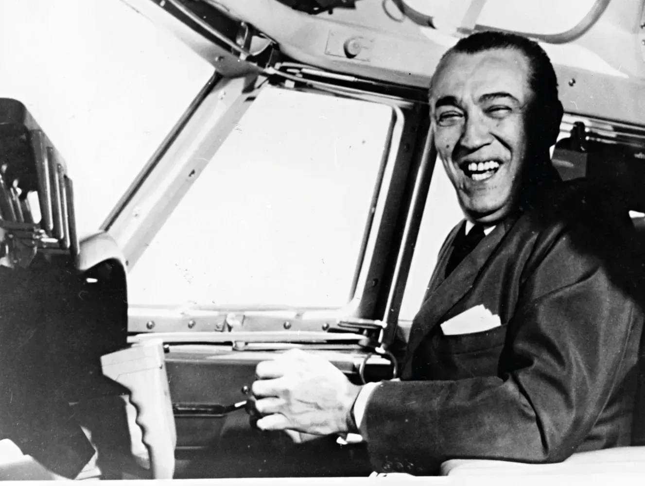 Uma pessoa sorridente, vestindo terno, está sentada na cabine de comando de um avião, segurando o manche.