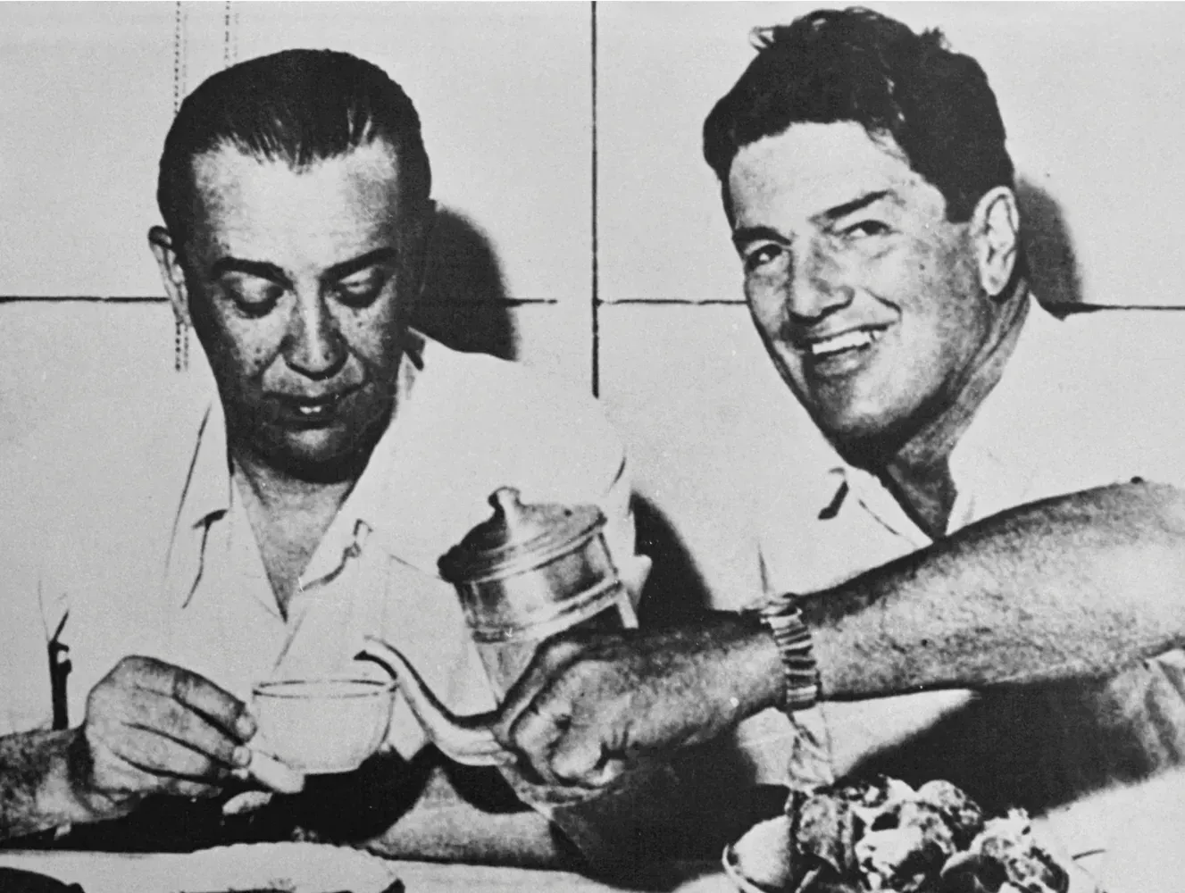 Dois homens sentados à mesa; um deles está servindo uma bebida no copo do outro enquanto sorri.