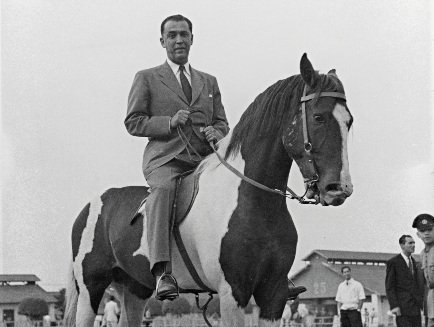 Um homem de terno monta um cavalo preto e branco ao ar livre, com observadores ao fundo.