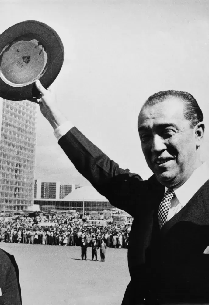 Juscelino Kubitschek acena com o chapéu para uma multidão em Brasília, com os prédios do Congresso visíveis ao fundo.