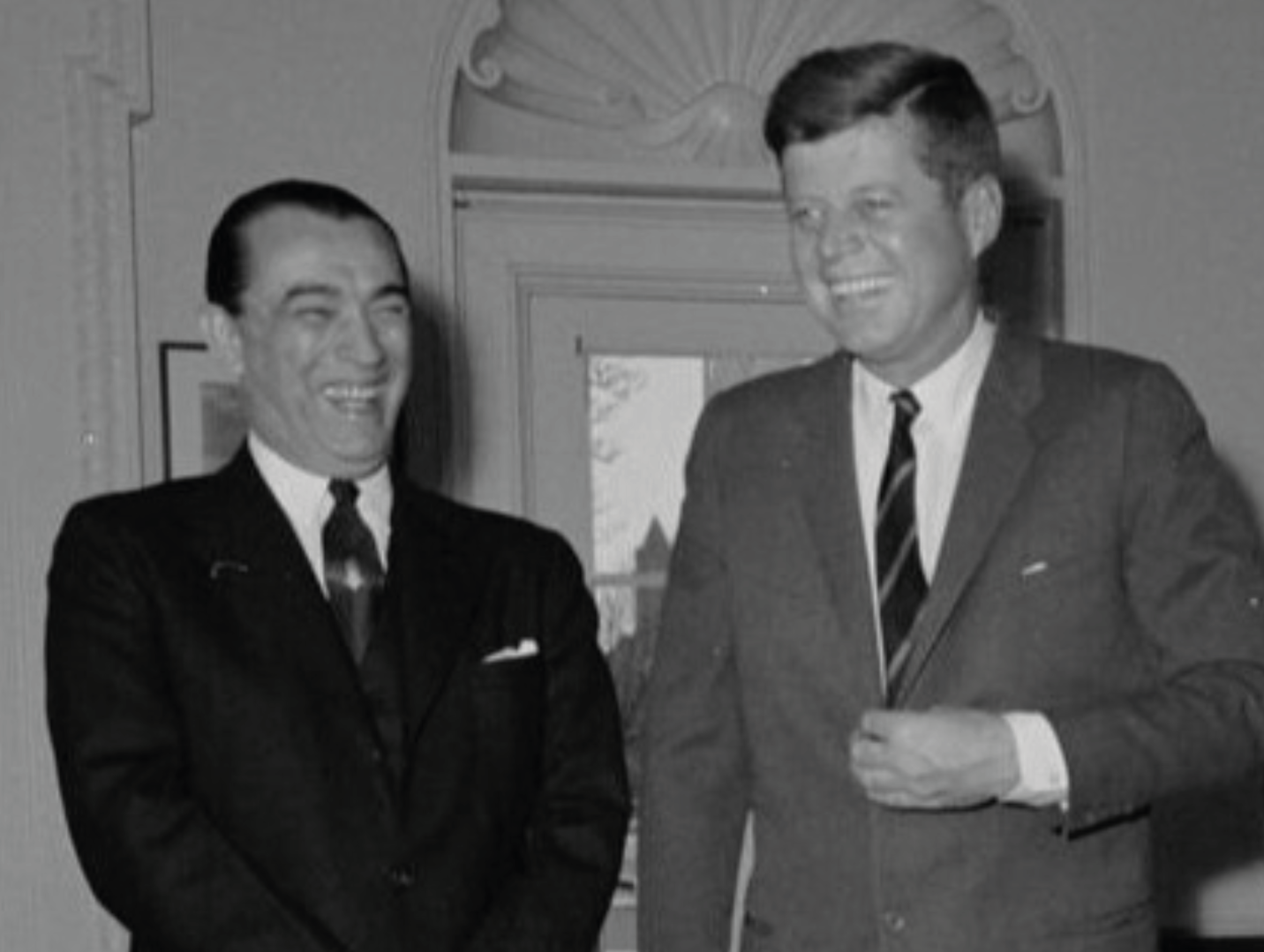 John F. Kennedy está ao lado de um homem, ambos sorrindo, de terno e gravata, em um ambiente interno.