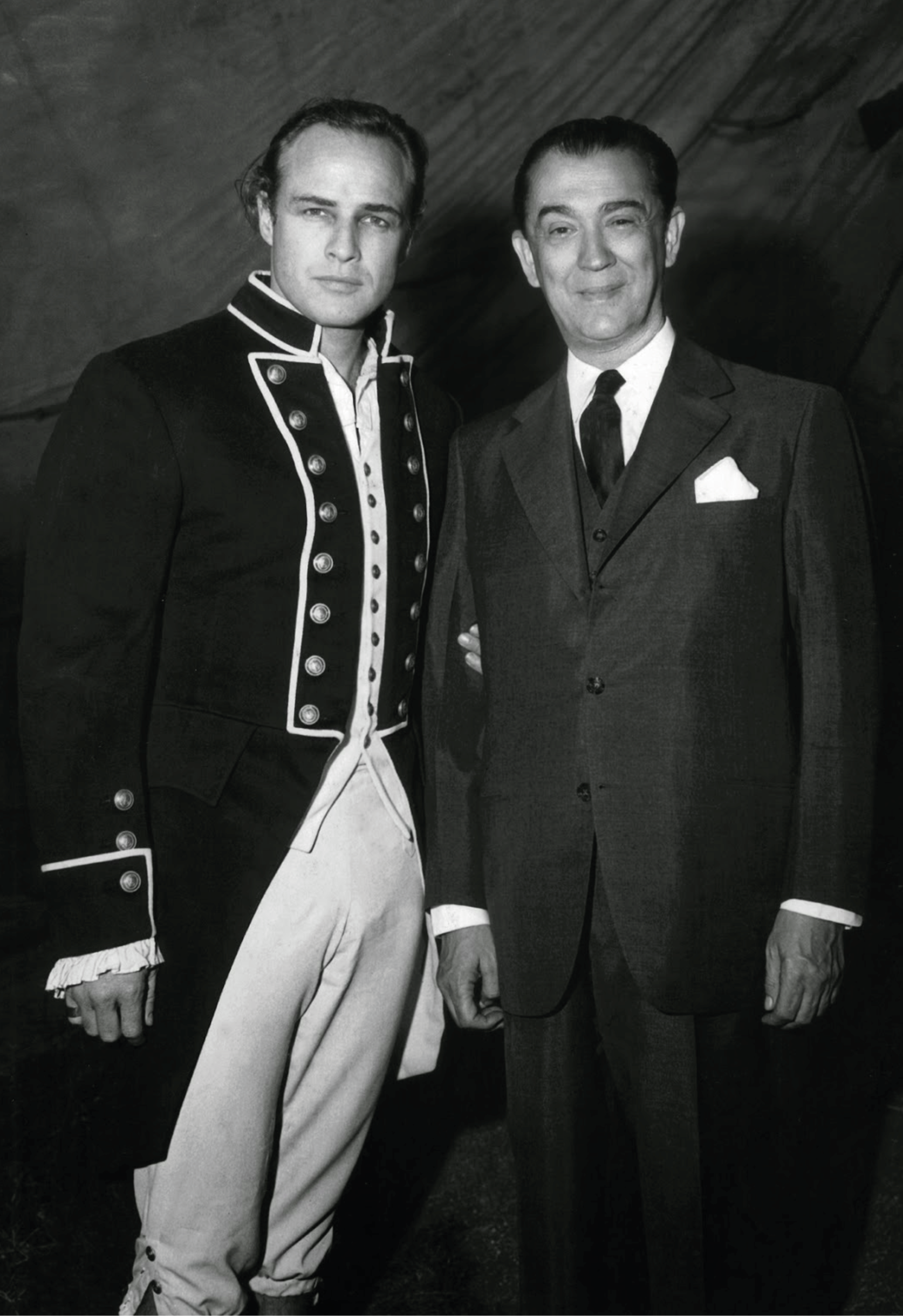 Marlon Brando em uniforme naval do século XVIII ao lado de um homem de terno e gravata.