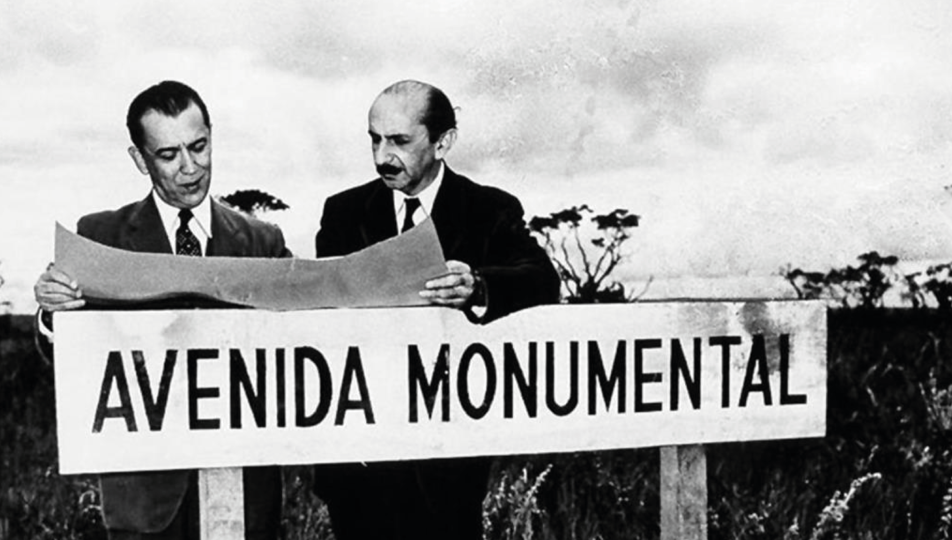 Dois homens de terno examinam plantas arquitetônicas atrás de uma placa de madeira com a inscrição