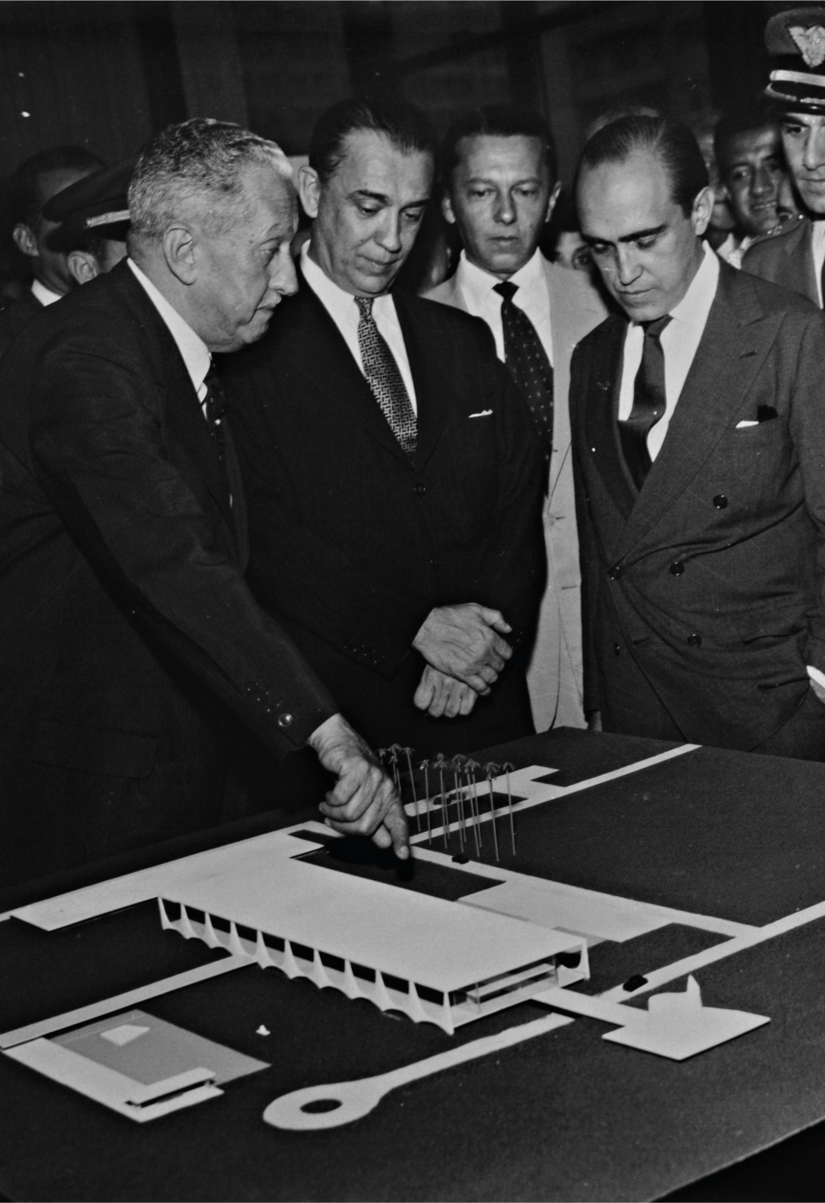 Quatro homens de terno estão em volta de uma maquete arquitetônica, com um deles gesticulando em direção a ela, em um ambiente formal e profissional.