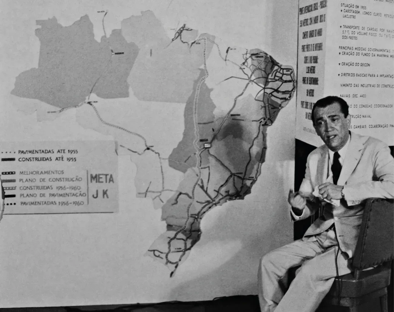 Juscelino Kubitschek aponta para um mapa do Brasil detalhando seu plano de infraestrutura e construção de estradas