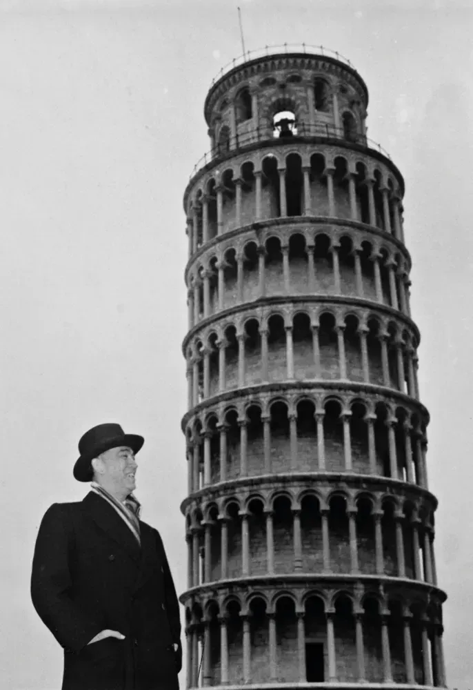 Uma pessoa de chapéu e casaco está em frente à Torre Inclinada de Pisa, olhando para a estrutura.