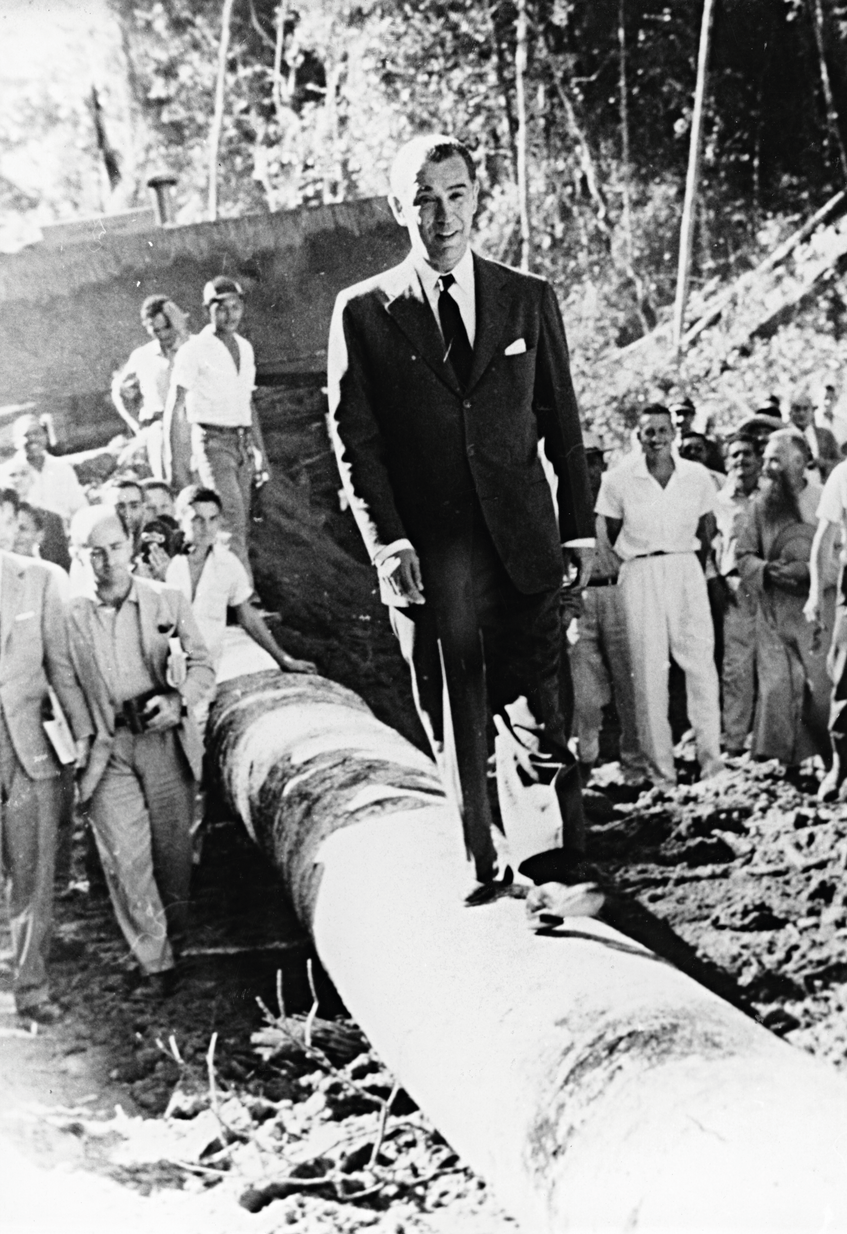 Um homem de terno atravessa um grande cano em um canteiro de obras industrial, cercado por observadores.
