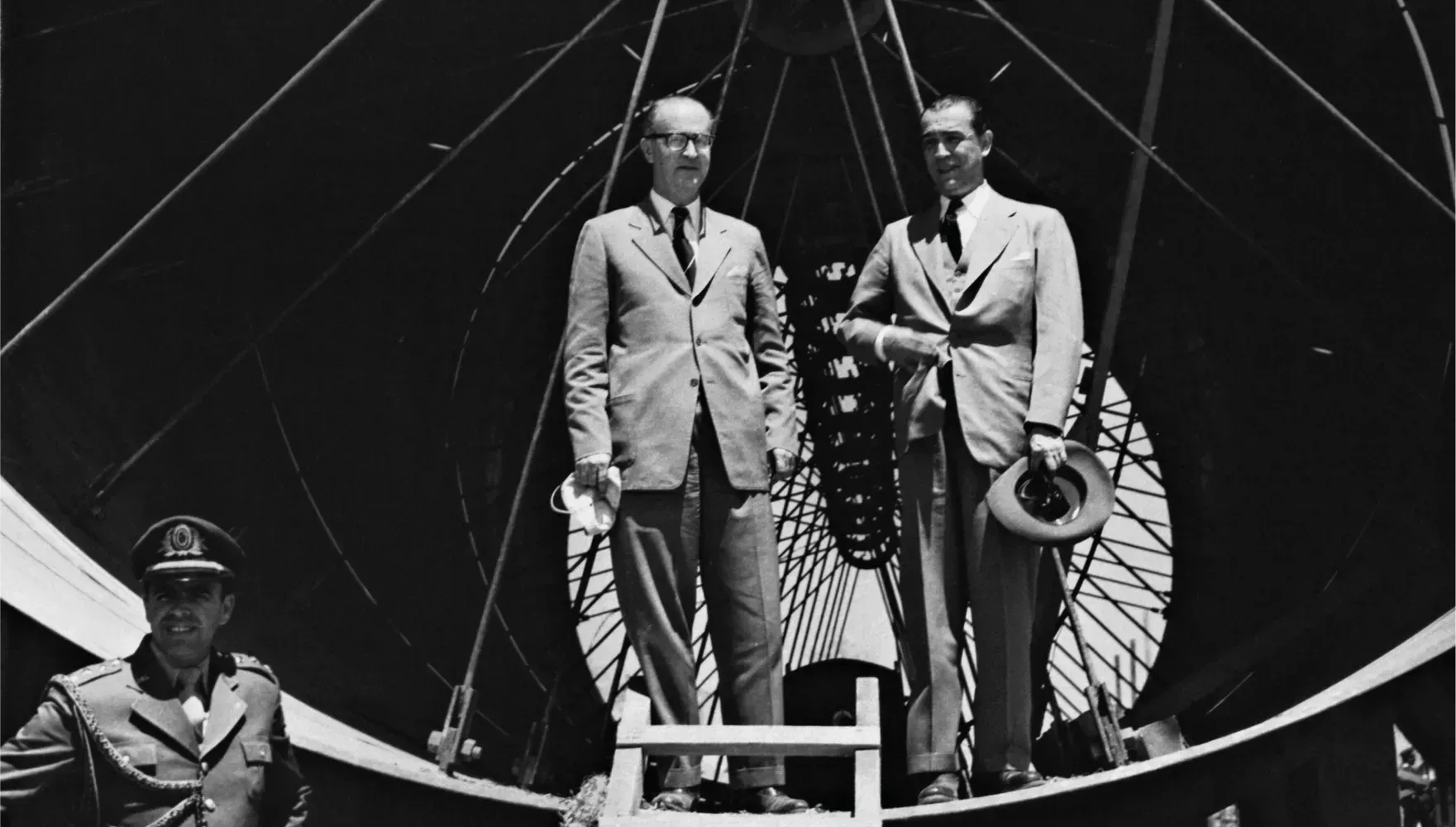 Dois homens de terno e um oficial uniformizado estão de pé diante da estrutura circular interna de um dirigível ou de uma grande aeronave.
