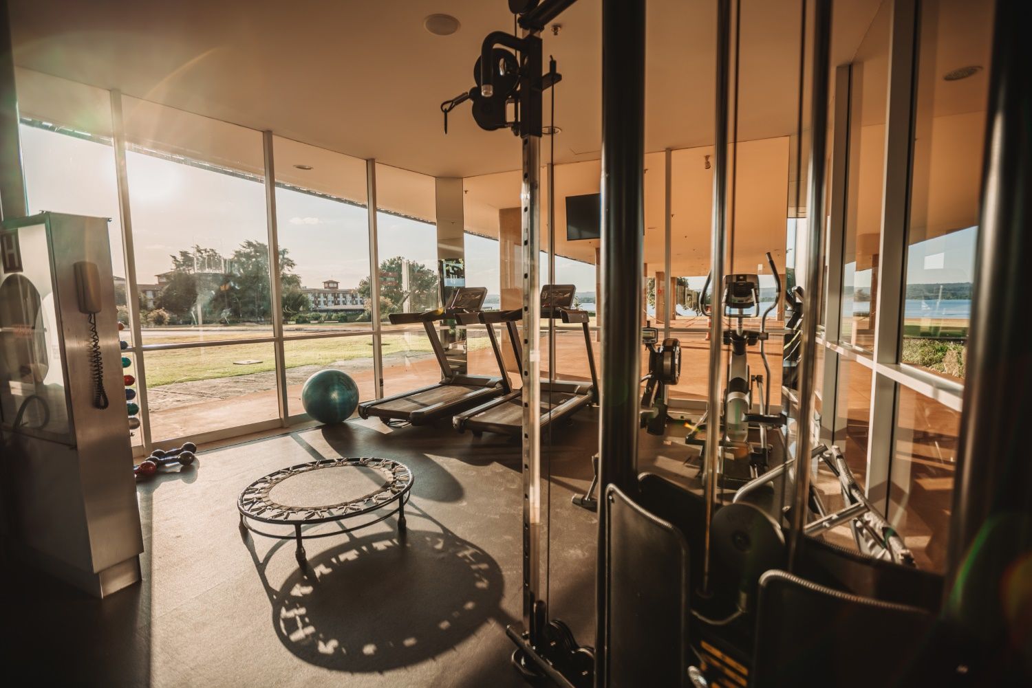 Academia com diversos equipamentos de ginástica, janelas amplas e vista para o exterior.