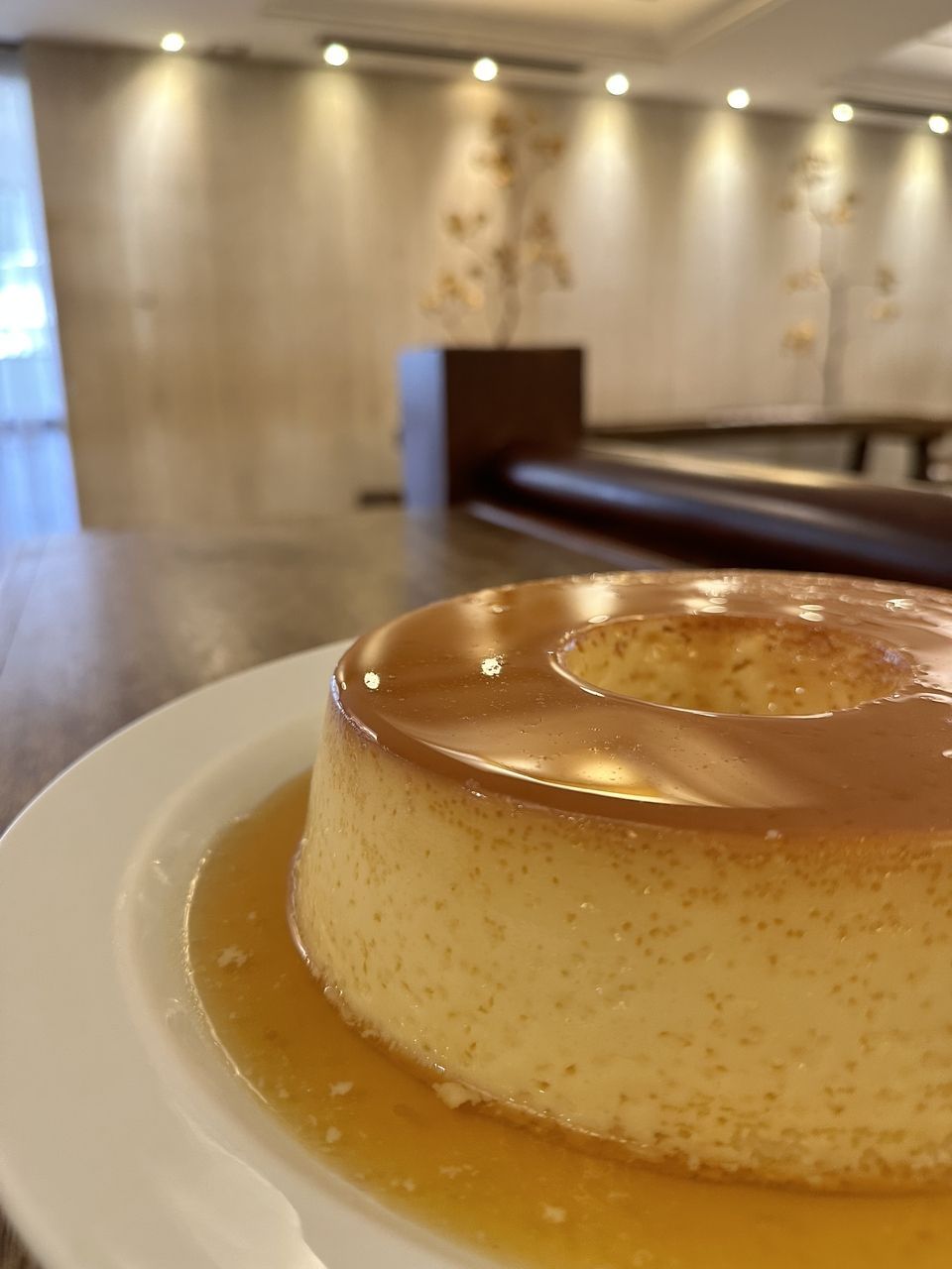 Pudim em um prato branco, com calda de caramelo, em uma sala de jantar.